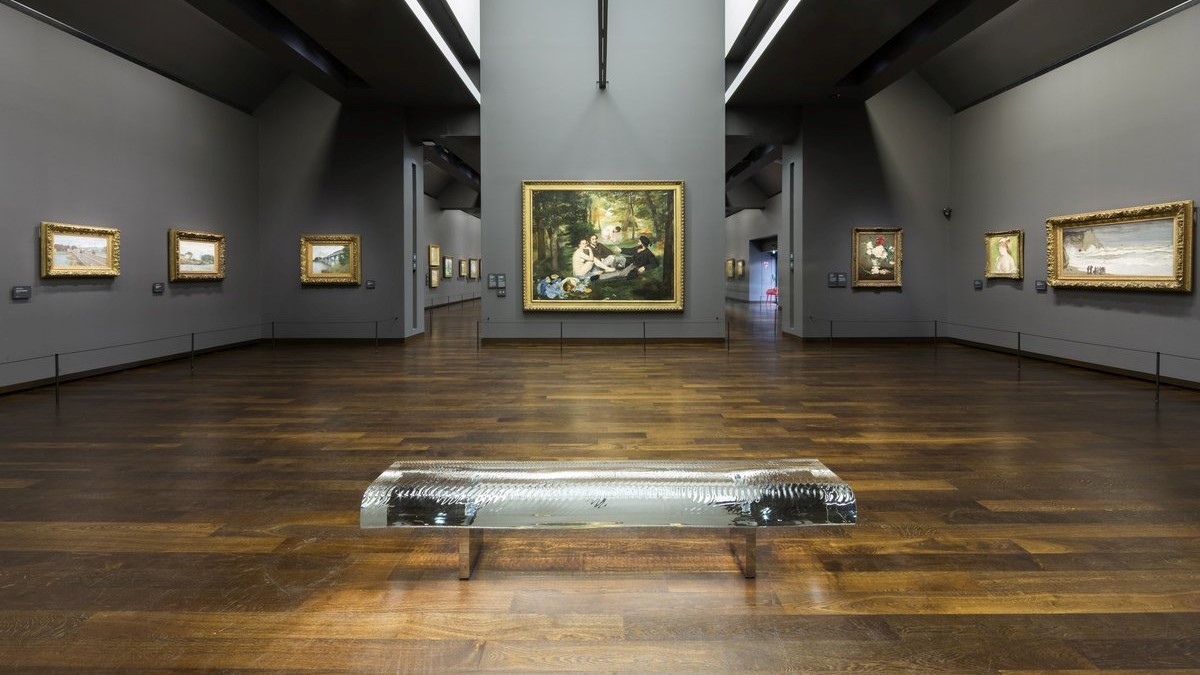 Large room with wooden floor, one chair and grey walls filled with painting at Musee D'Orsay