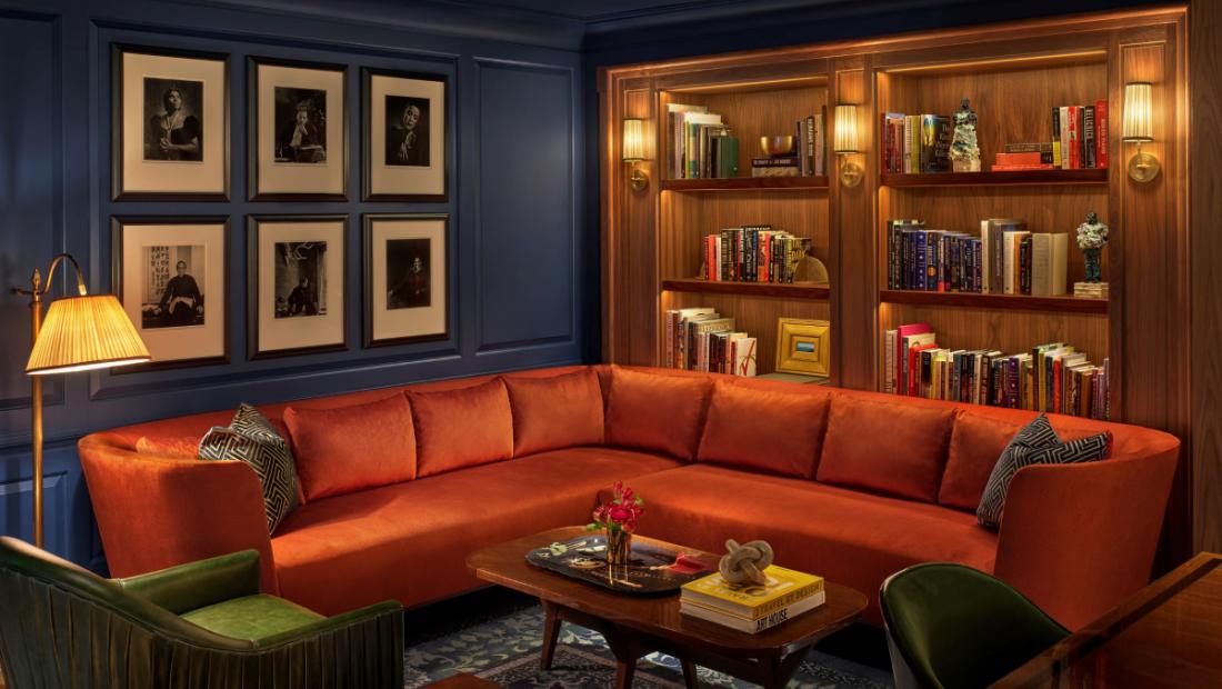 A lounge/tea area with a red sofa, blue squared walls with six paintings and a brown bookshelf.