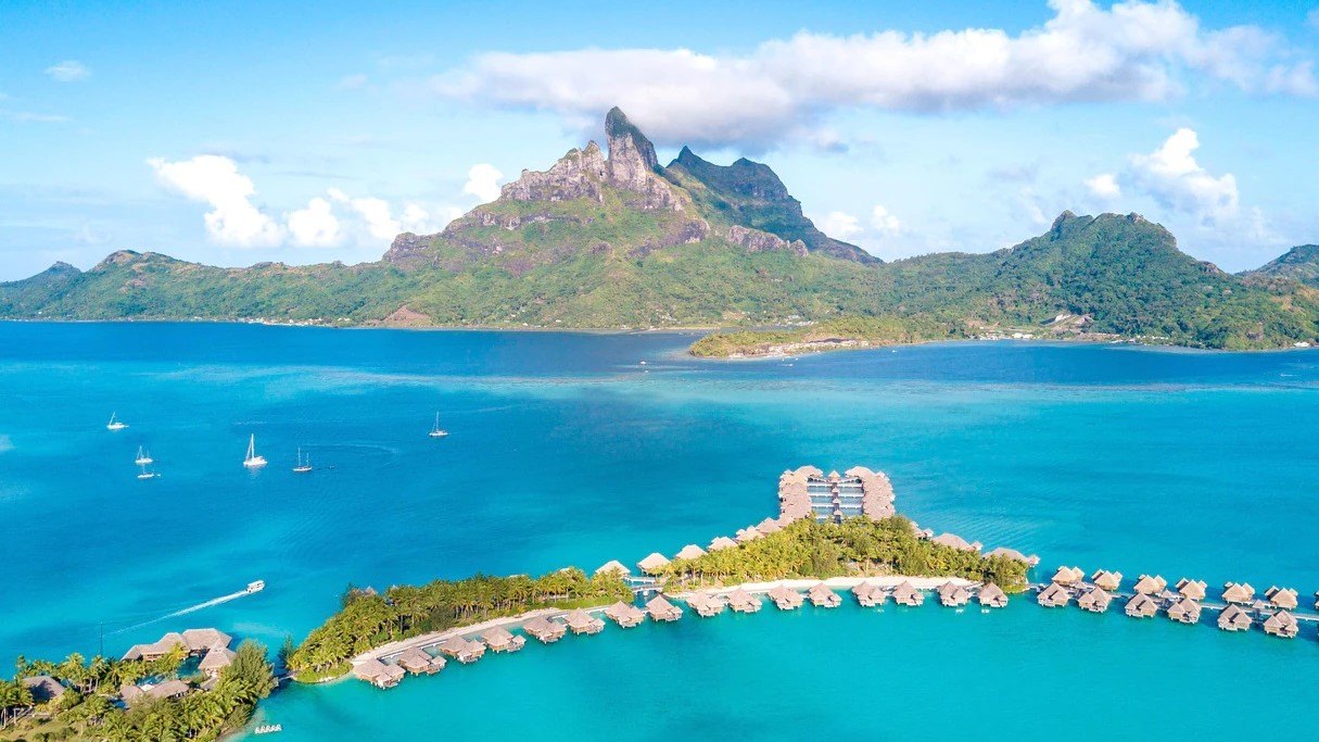 Bird view of The St Regis Bora Bora Resort with clear water, nature and mountains not far away, petite island with cabins and nature, and sailing boats in white passing by.