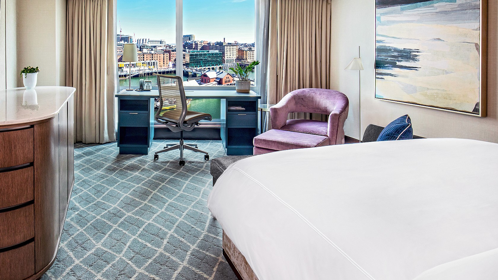 Hotel room with a bed, a blue desk, a pink chair and windows with view over the city. 