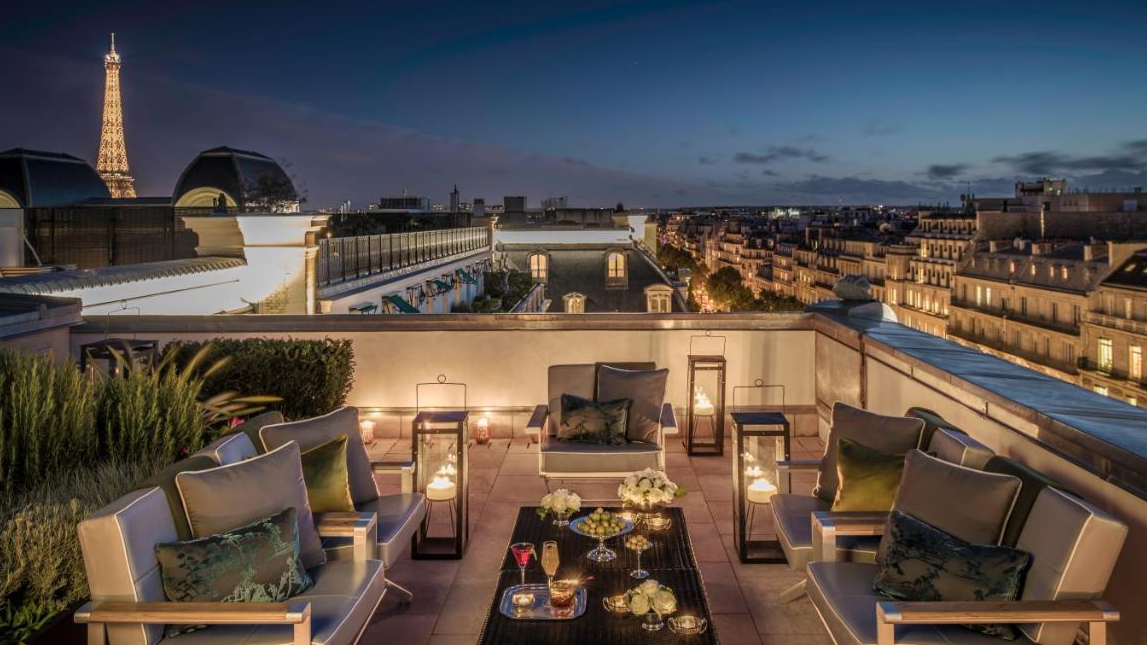 Rooftop bar in Paris with view of the Eiffel Tower, olives, and drinks at the table surrounded by plants, grey furniture and tender lightning at Le Rooftop - Hotel Peninsuala Paris.