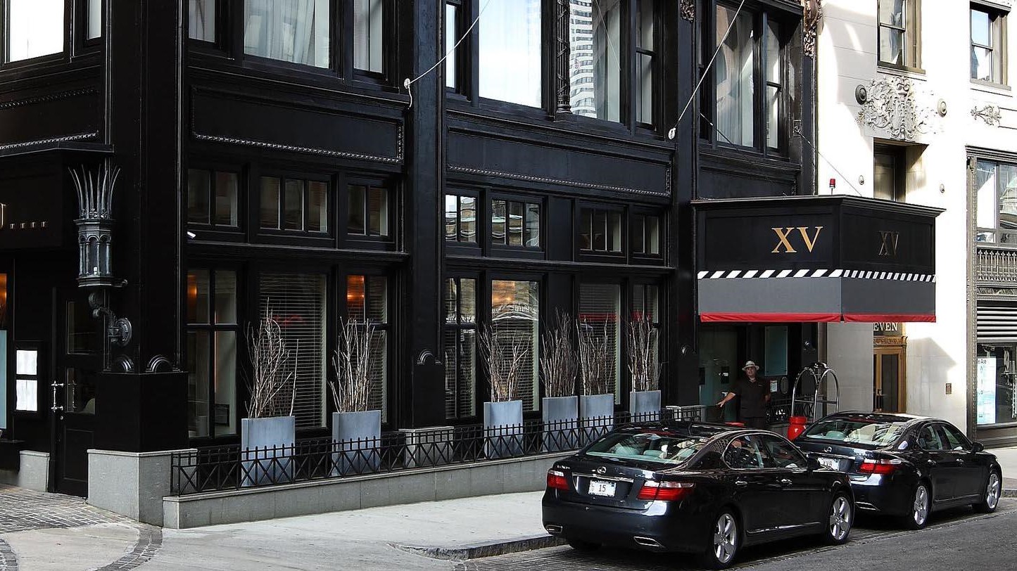 The outside of a all black hotel XV beacon with XV written in gold and two black mercedes parked upfront.