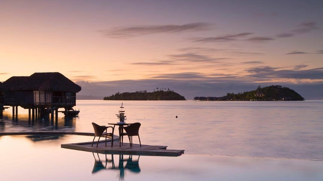 Small water bridge to a private seating area, wooden cabins and water and sun setting.